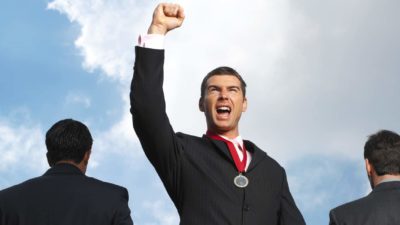 A businessman in a suit wears a medal around his neck and raises a fist in victory surrounded by two other businessmen in suits facing the other direction to him.