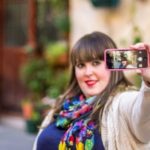 A woman in colourful outfit holds up a phone to take a selfie.