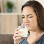 A woman in her 20s holds a glass of milk up towards her face as if to drink it but makes a grimacing face as though she has smelt that the milk might be off or soured.