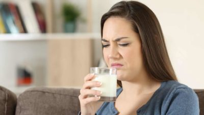 A woman in her 20s holds a glass of milk up towards her face as if to drink it but makes a grimacing face as though she has smelt that the milk might be off or soured.