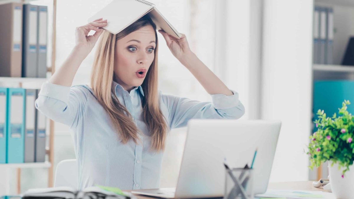 A young woman holds an open book over her head with a round mouthed expression as if to say oops as she looks at her computer screen in a home office setting with a plant on the desk and shelves of books in the background.