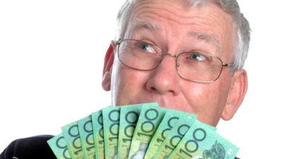 A mature aged man with grey hair and glasses holds a fan of Australian hundred dollar bills up against his mouth and looks skywards with his eyes as though he is thinking what he might do with the cash.