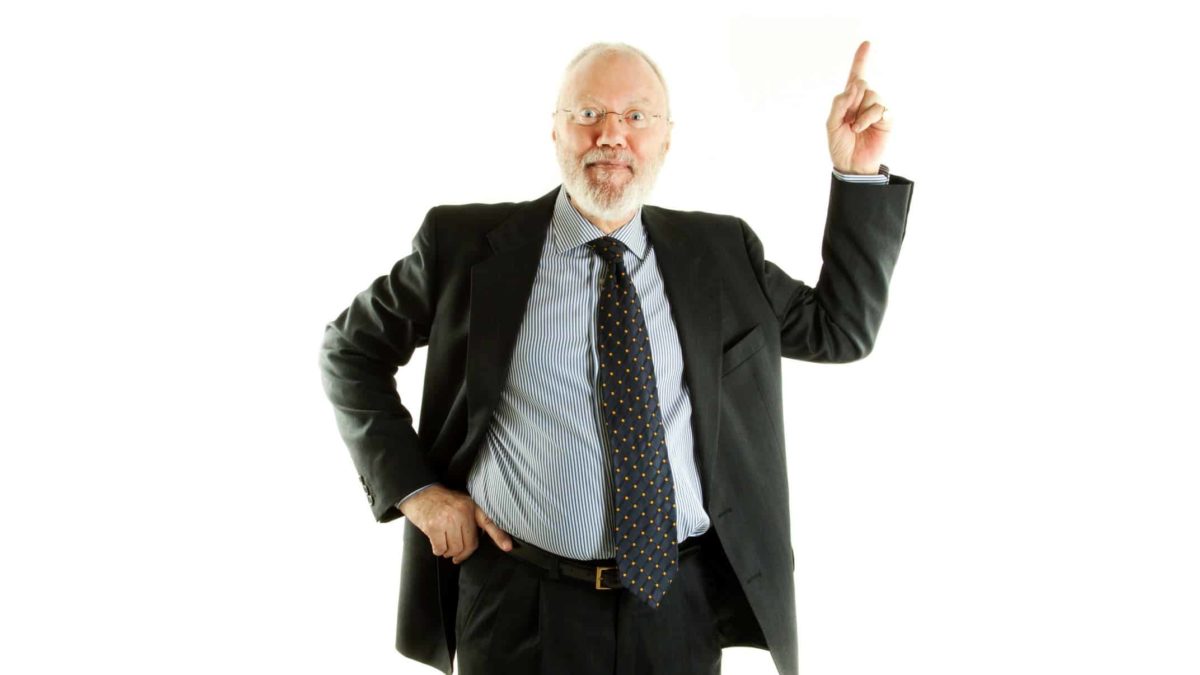 A middle aged businessman in a suit holds up one finger with his other hand on his hip with an enthusiastic, comical expression on his face.