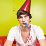A male party goer sits wearing a party hat and with a party blower in his mouth amid a bunch of balloons with a sad, serious look on his face as though the party is over or a celebration has fallen flat.