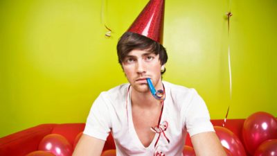 A male party goer sits wearing a party hat and with a party blower in his mouth amid a bunch of balloons with a sad, serious look on his face as though the party is over or a celebration has fallen flat.