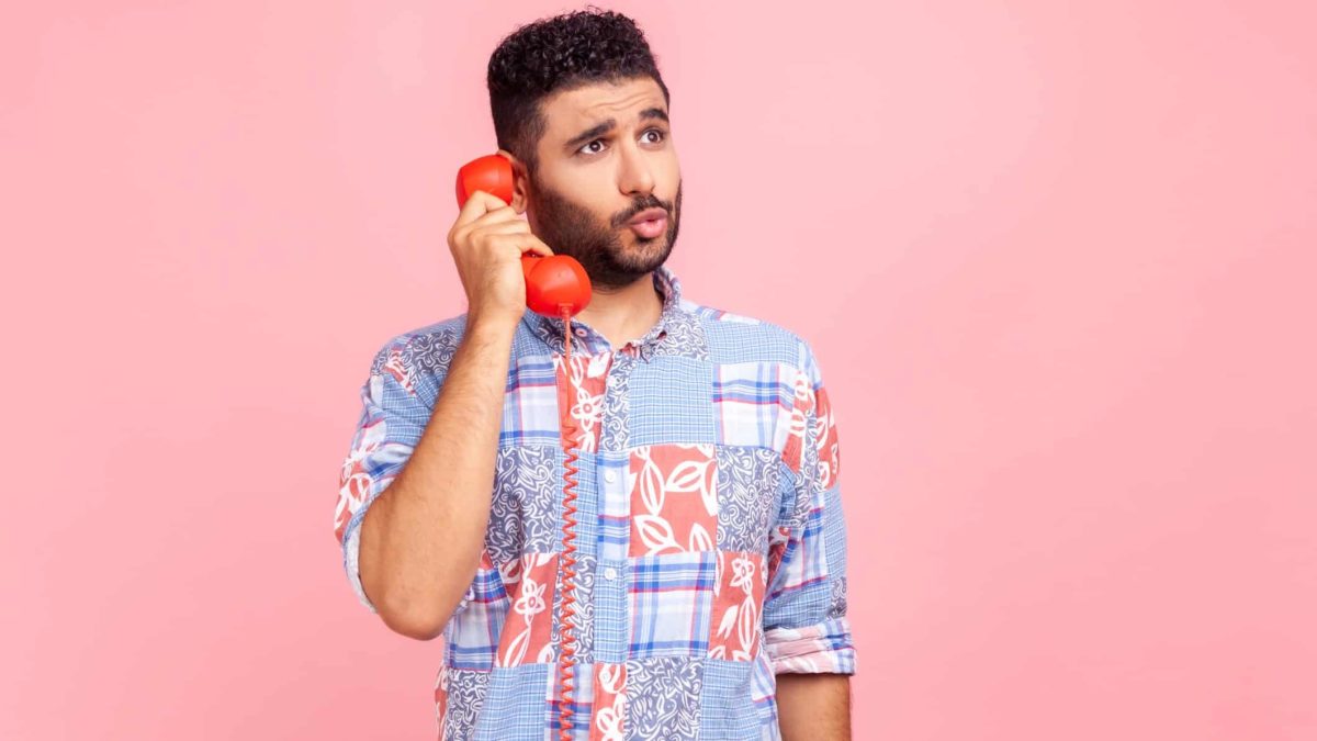 A man wearing a colourful shirt holds an old fashioned phone to his ear with a look of curiosity on his face as though he is pondering the answer to a question.