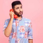 A man wearing a colourful shirt holds an old fashioned phone to his ear with a look of curiosity on his face as though he is pondering the answer to a question.