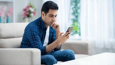 A man sits in contemplation on his sofa looking at his phone as though he has just heard some serious or interesting news.