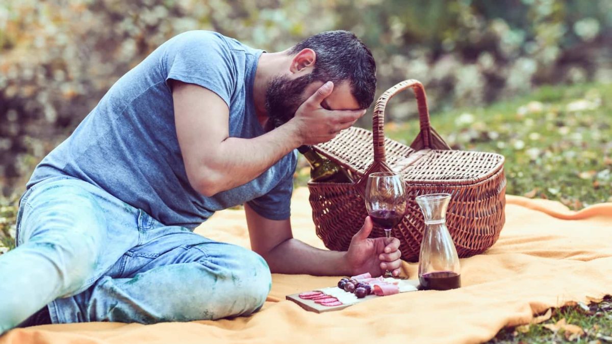 A man puts his head in his hand as he sits on rig with picnic basket and wine.