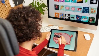 A child photographed from behind draws a cute picture of a pig on a digital screen with another larger screen on a desk in front of him filled with multiple images of similar cartoon style pictures.