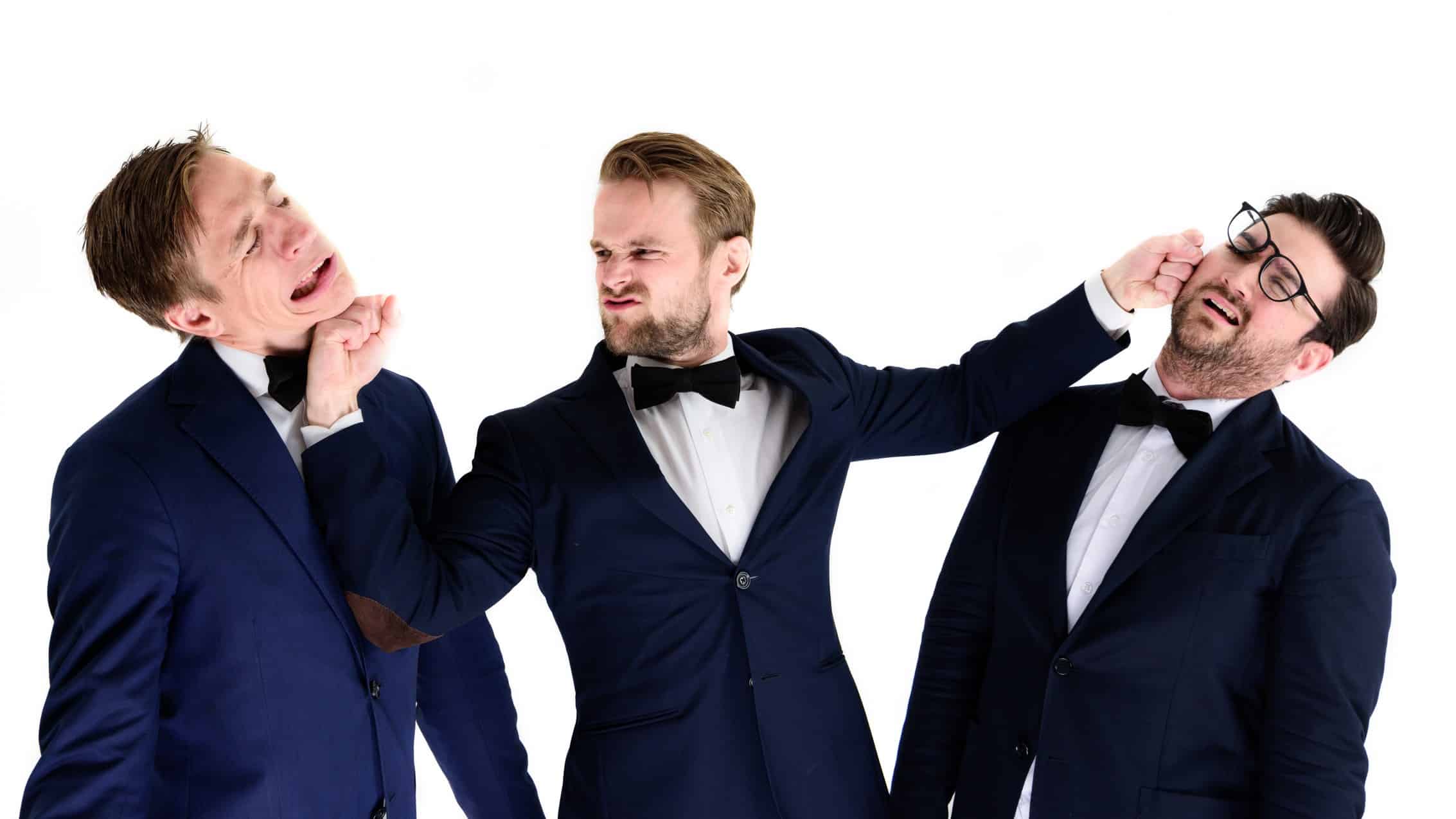 A group of three young men in dinner suits lark around with the one in the middle pretending to deliver blows to the faces of his companions while they make exaggerated expressions of pain and suffering.