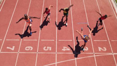 Five people are lunging for the finish line on an athletics track with the picture taken from above as an aerial view of the athletes with their arms outstretched.