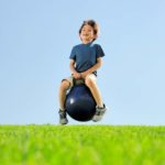 A young boy sits on top of a big rubber bouncing ball with handles as he smiles a toothless grin at the camera and bounces above the ground in a grassy field with a blue sky.