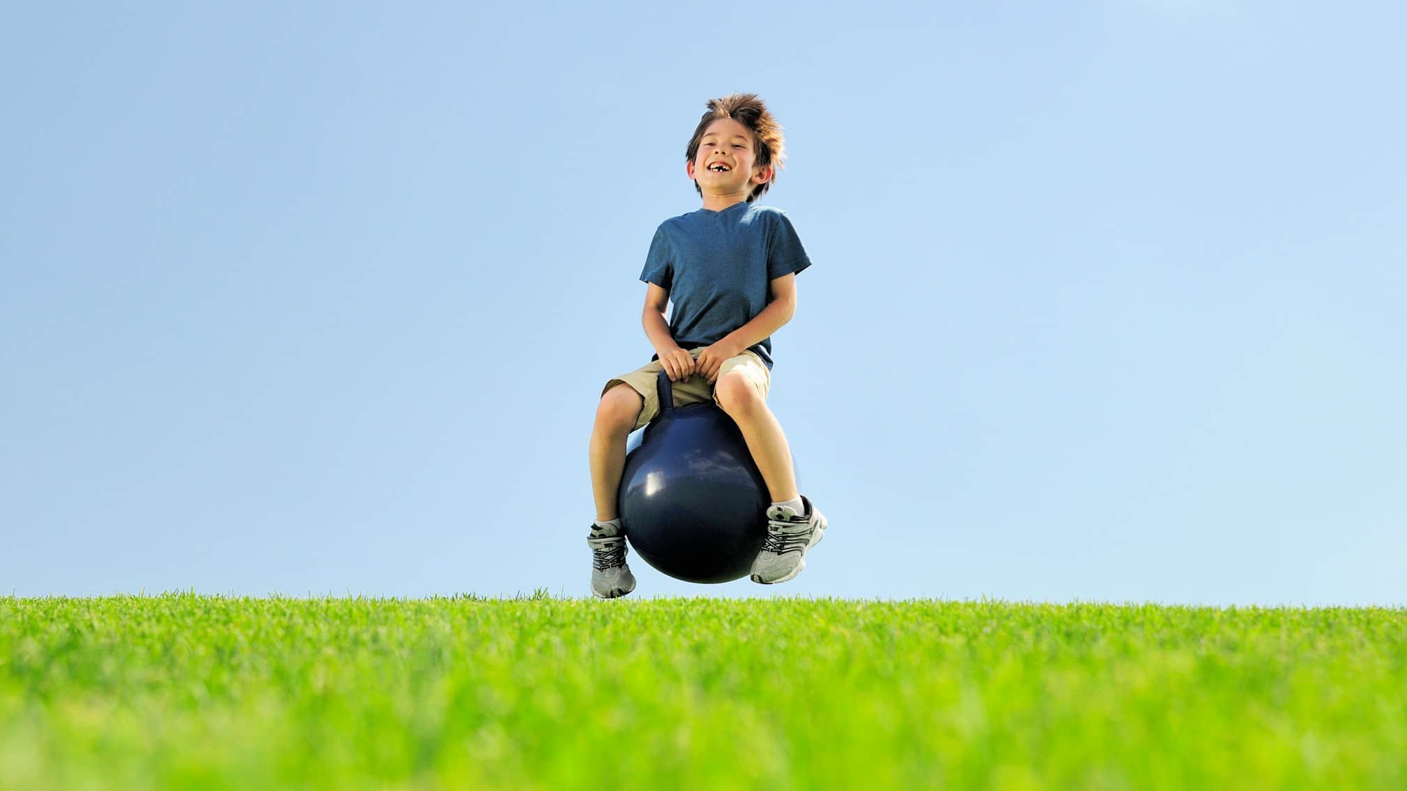 A young boy sits on top of a big rubber bouncing ball with handles as he smiles a toothless grin at the camera and bounces above the ground in a grassy field with a blue sky.