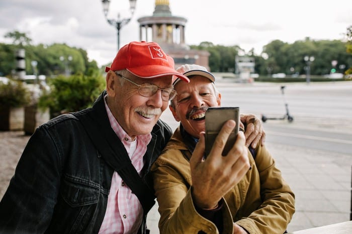 Two elderly men laugh together as they take a selfie with a mobile phone with a city scape in the background.