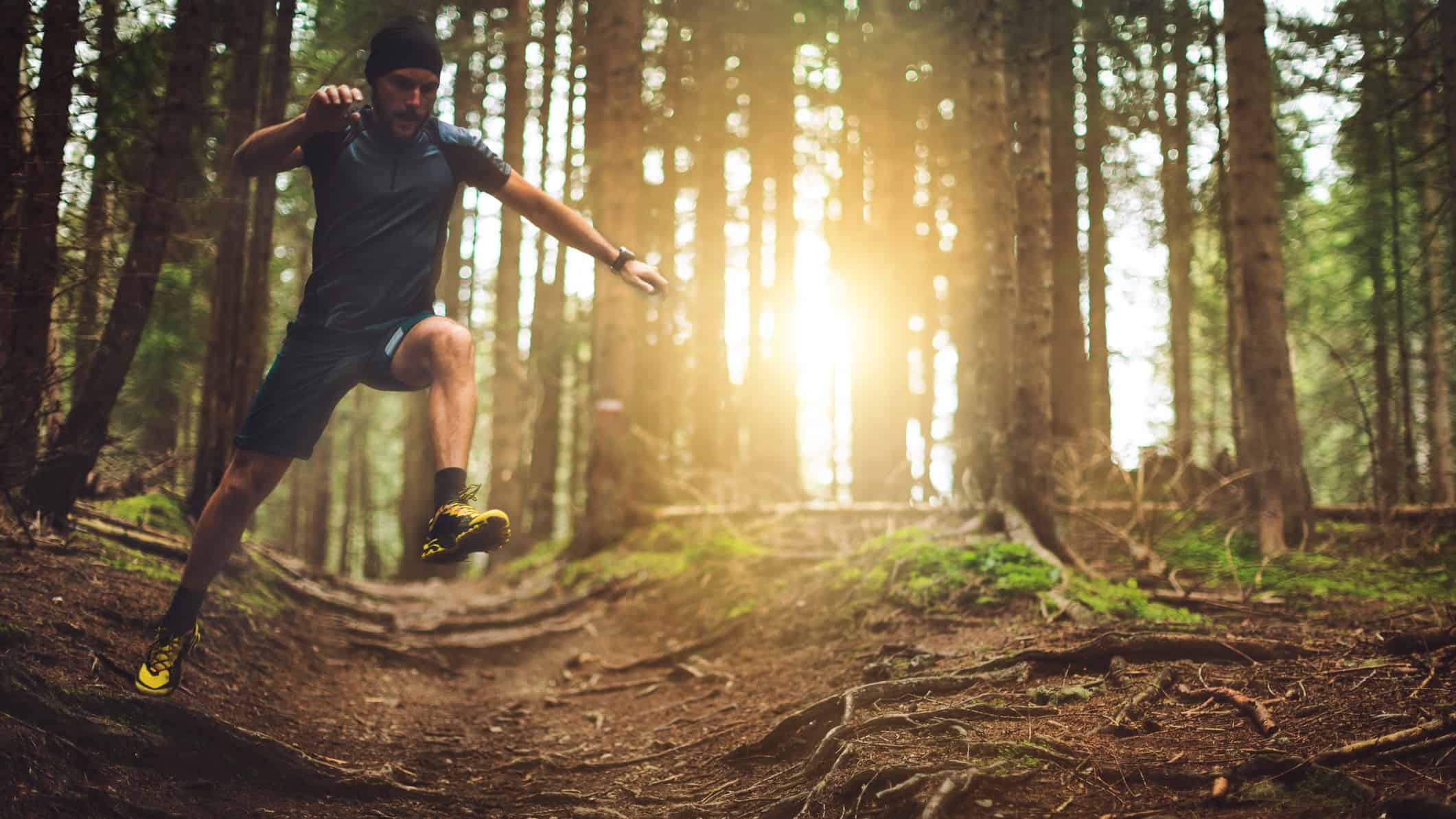 A male runner is in an awkward pose as he approaches an uneven part of a running track through a forest with tall trees and sunlight shining through them.