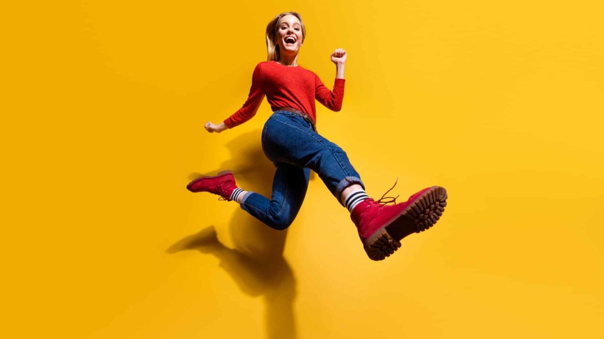 A young woman dressed in street clothes leaps happily in the air with the focus on her bright red boots that are front and centre for the camera.
