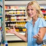 A frustrated young woman shopper holds her hands up with a pained, annoyed expression on her face as she stands next to her trolley in a grocery store and examines the stock offerings on the shelf in front of her.
