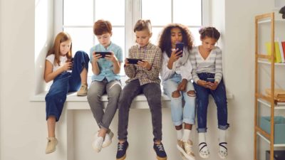 A group of young kids, aged 12-13, sit together side by side on a window ledge with all looking at their mobile phones in their hands with sombre, serious expressions on their faces as if they are engaged in social media.