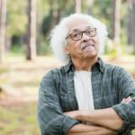 An old man with wavy white hair folds his arms in a stubborn gesture as he stands defiantly in an outdoor setting.