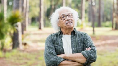 An old man with wavy white hair folds his arms in a stubborn gesture as he stands defiantly in an outdoor setting.