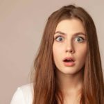 A young woman with long brown hair opens her green eyes and mouth widely, expressing surprise.