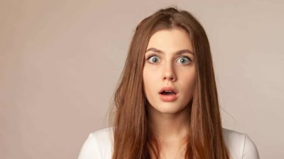 A young woman with long brown hair opens her green eyes widely expressing surprise.