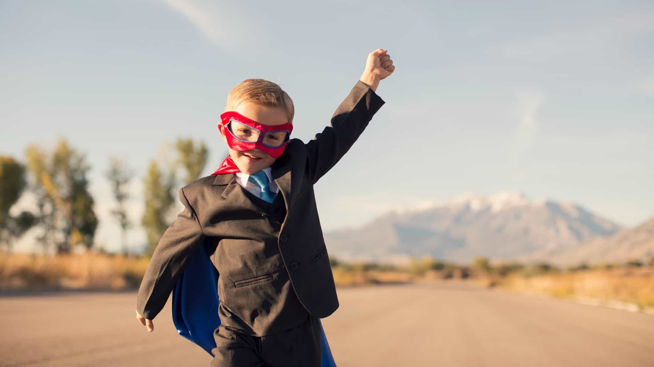 A small child dressed in a business suit and a superhero mask and cape holds a hand aloft in a superhero pose against the background of a barren, dusty landscape.