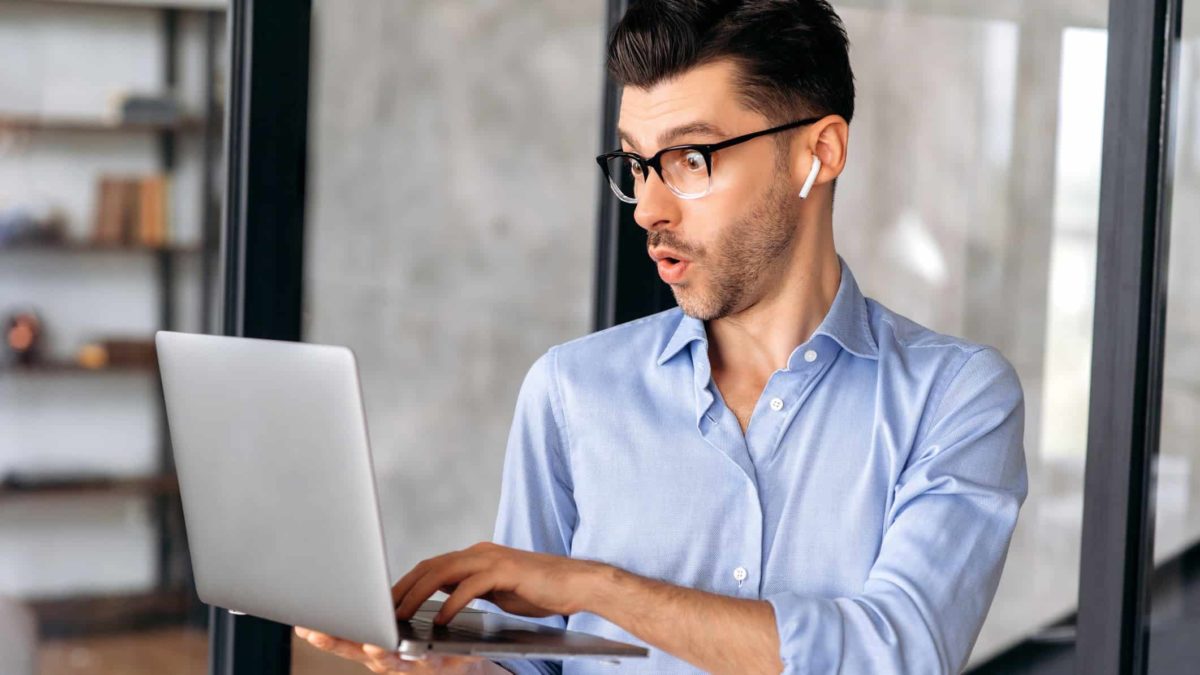 A man in his 30s holds his laptop and operates it with his other hand as he has a look of pleasant surprise on his face as though he is learning something new or finding hidden value in something on the screen.