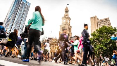 A scene of a busy city streeet in Sydney with an old town hall in the background next to a modern high rise building with a blur of people walking in multiple directions on the footpaths in front of them.