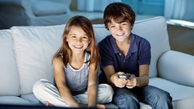 Two cute young children, a boy and a girl, sit on a sofa together with eager looks on their faces as the boy holds a remote control in one hand.