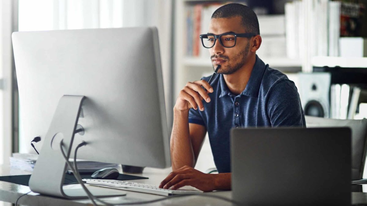 A man sits in deep thought with a pen held to his lips as he ponders his computer screen with a laptop open next to him on his desk in a home office environment.