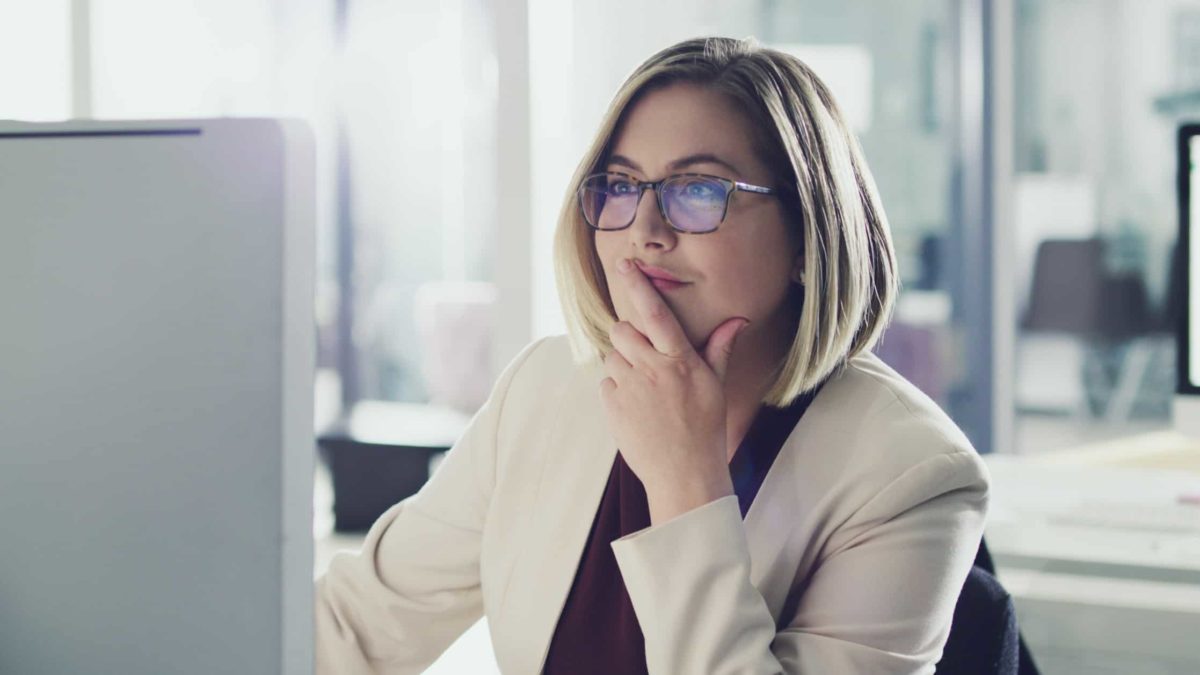 A woman sits at her computer with her hand to her mouth and a contemplative smile on her face as she reads about the performance of Allkem shares on her computer