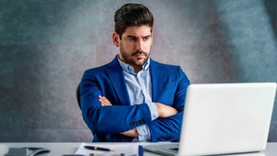 An unhappy man in a suit sits at his desk with his arms crossed staring at his laptop screen as the PointsBet share price falls