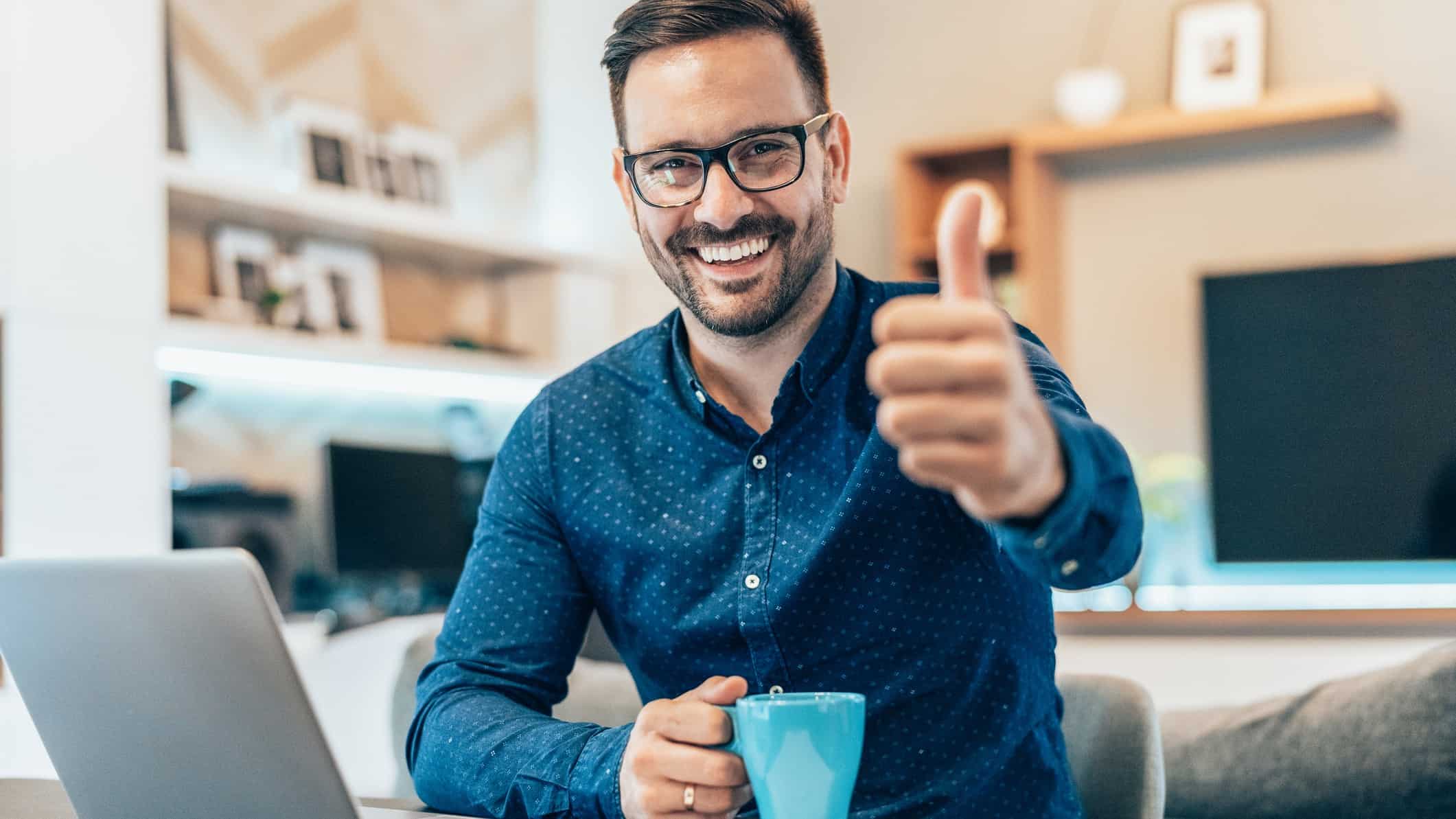 A man holding cup of coffee puts his thumb up and smiles while at laptop.