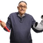 A middle aged man holds a plumbing plunger in one hand and a piece of toilet pipe in the other, with an exasperated look on his face.