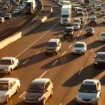 Many cars travell on a busy six lane road way with other cars in the background travelling in the opposite direction, going the other way.dway