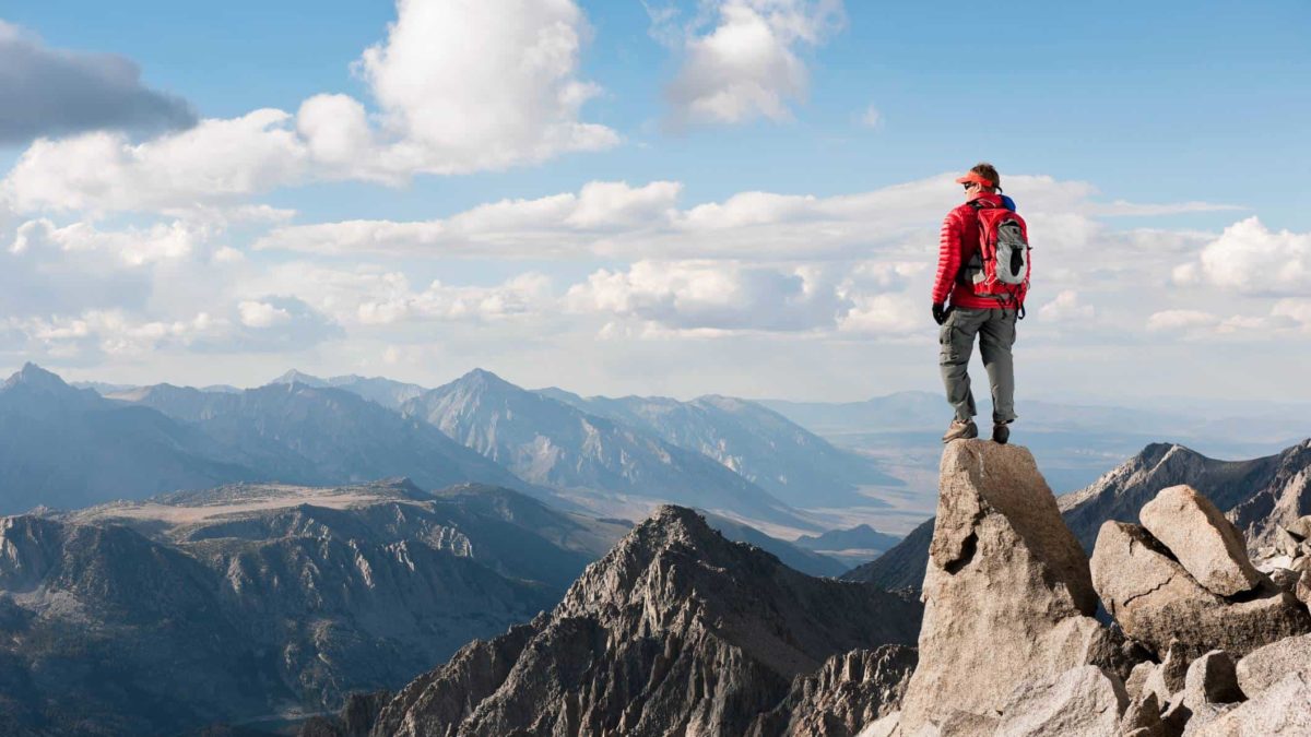 A man wearing a red jacket and mountain hiking clothes stands at the top of a mountain peak and looks out over countless mountain ranges.