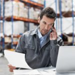 A warehouse storeman sits in front of a computer with a phone to his ear and paper in one hand with a well stocked warehouse in the background.