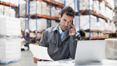 A warehouse storeman sits in front of a computer with a phone to his ear and paper in one hand with a well stocked warehouse in the background.