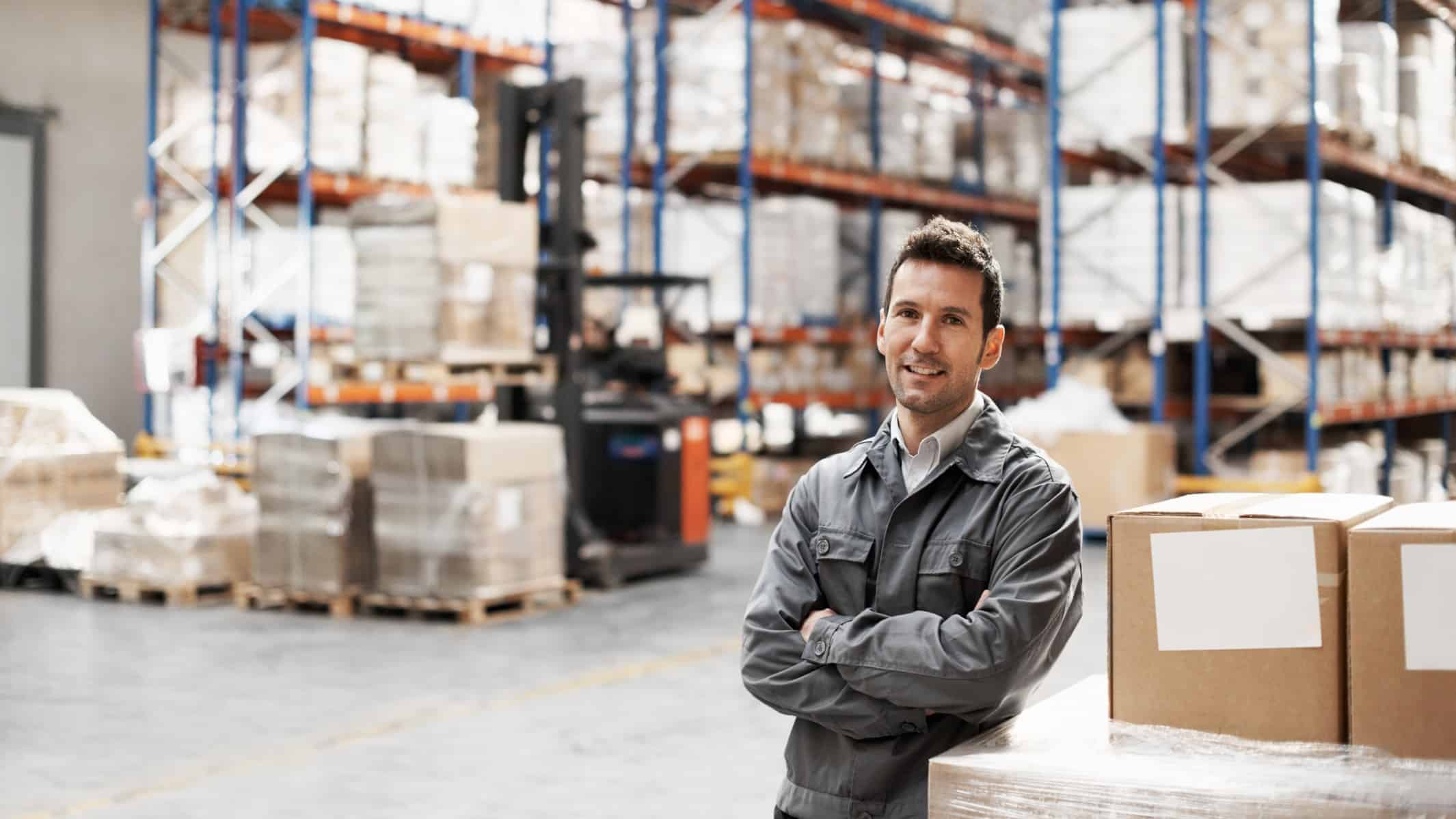 A warehouse storeman stands with his arms folded in front of a well-stocked warehouse interior with goods and moving equipment in the background.