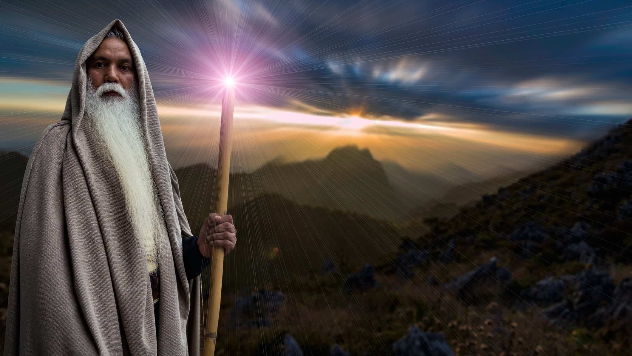 An old man wearing long robes with a long white beard holds a staff that emits light in all directions as he stands in front of a mystical landscape of mountains and a rising sun.