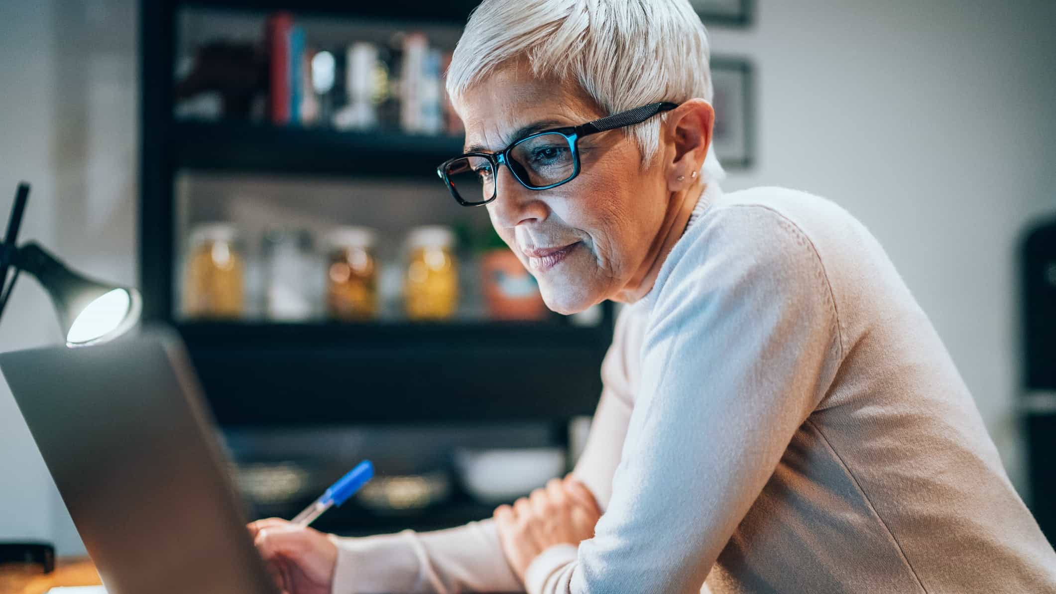 A mature age woman with a groovy short haircut and glasses, sits at her computer, pen in hand thinking about information she is seeing on the screen.