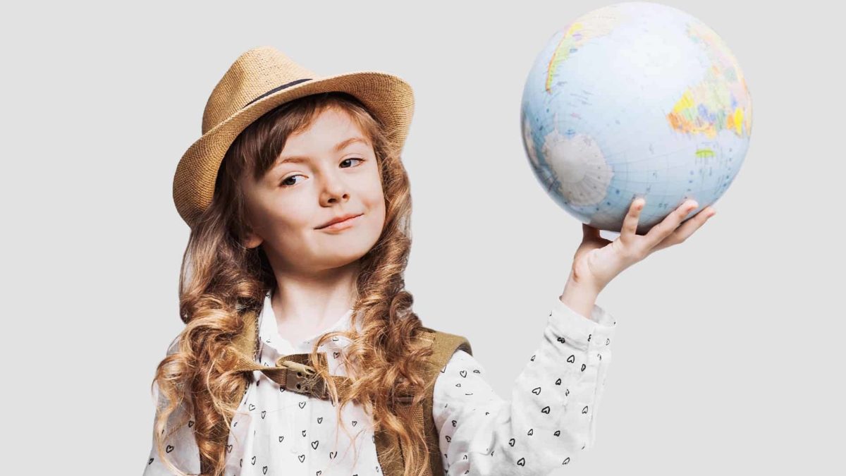 A cute young girl wears a straw hat and has a backpack strapped on her back as she holds a globe in her hand with a cheeky smile on her face.