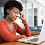 A woman sits at a computer with a quizzical look on her face with eyerows raised while looking into a computer, as though she is resigned to some not pleasing news.