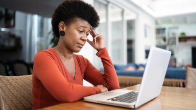 A woman sits at a computer with a quizzical look on her face with eyerows raised while looking into a computer, as though she is resigned to some not pleasing news.