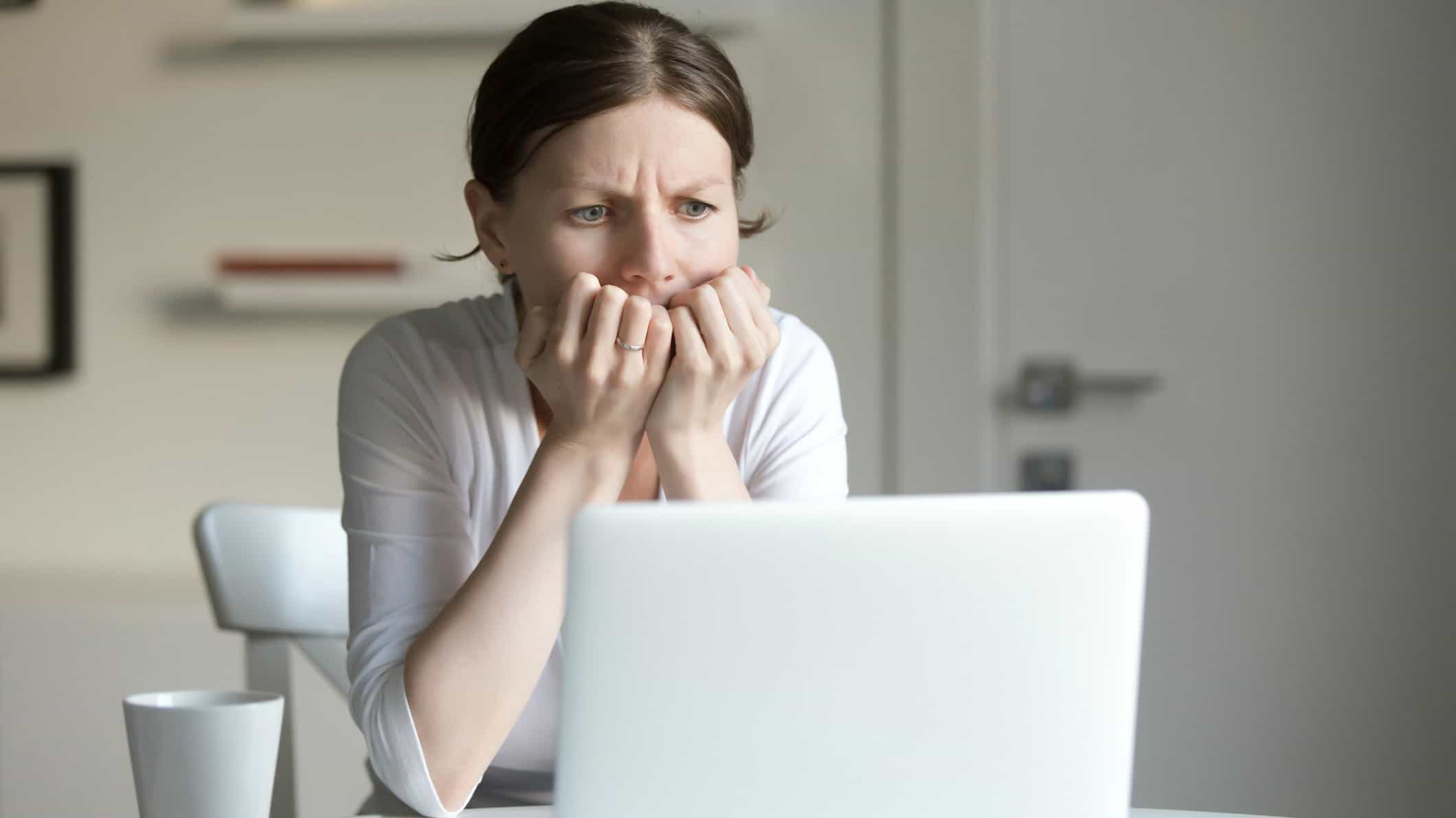 A woman sits at her computer with her hands clutched her the bottom of her face as though she may be biting her fingermails with a worried expression in her eyes and frown lines visible.