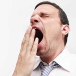 A businessman holds his hand to his wide-open yawning mouth as he closes his eyes and makes a funny face while he gives a wholehearted yawn.