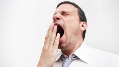 A businessman holds his hand to his wide-open yawning mouth as he closes his eyes and makes a funny face while he gives a wholehearted yawn.