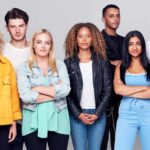A group of seven young people of different genders and cultural backgrounds stand in a group with serious expressions wearing casual young persons' attire.